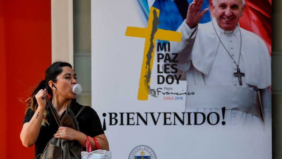 A woman blows a bubble with chewing gum, as she walks past a welcome poster with the image of Pope Francis, ahead of his upcoming visit to Santiago on January 11, 2018. Pope Francis will be visiting Chile from January 15 to 18. / AFP PHOTO / Martin BERNETTI (Photo credit should read MARTIN BERNETTI/AFP/Getty Images)