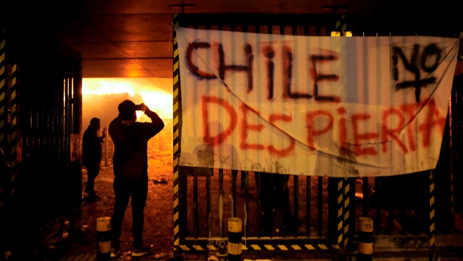 TOPSHOT - People take pictures of Macul Metro station set on fire by protesters alongside a sign that reads "Chile doesn't wake up" during a mass fare-dodging protest in Santiago, on October 19, 2019. - The entire Santiago Metro, which mobilizes about three million passengers per day, stopped operating on Friday afternoon following attacks in rejection of the rate hike, the company said. The chaos beat Santiago this Friday with confrontations, fires and attacks on the metropolitan railway, in protest of the increase in fares that forced the closure of all Metro stations. (Photo by JAVIER TORRES / AFP) (Photo by JAVIER TORRES/AFP via Getty Images)